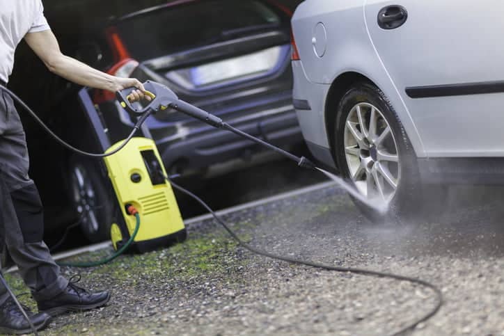 A person cleaning the car by a garage with a high pressure washer.